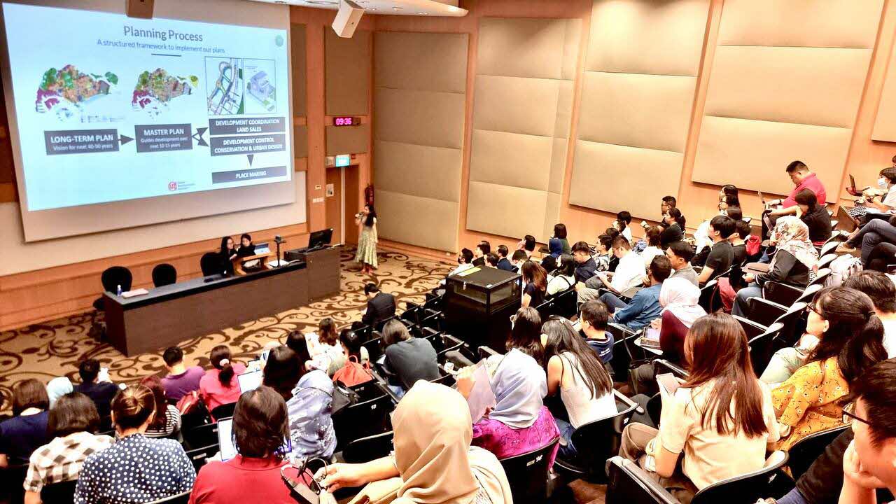 Lecture-style sharing session on urban planning, with participants seated in an auditorium watching a presentation on Singapore’s planning process.